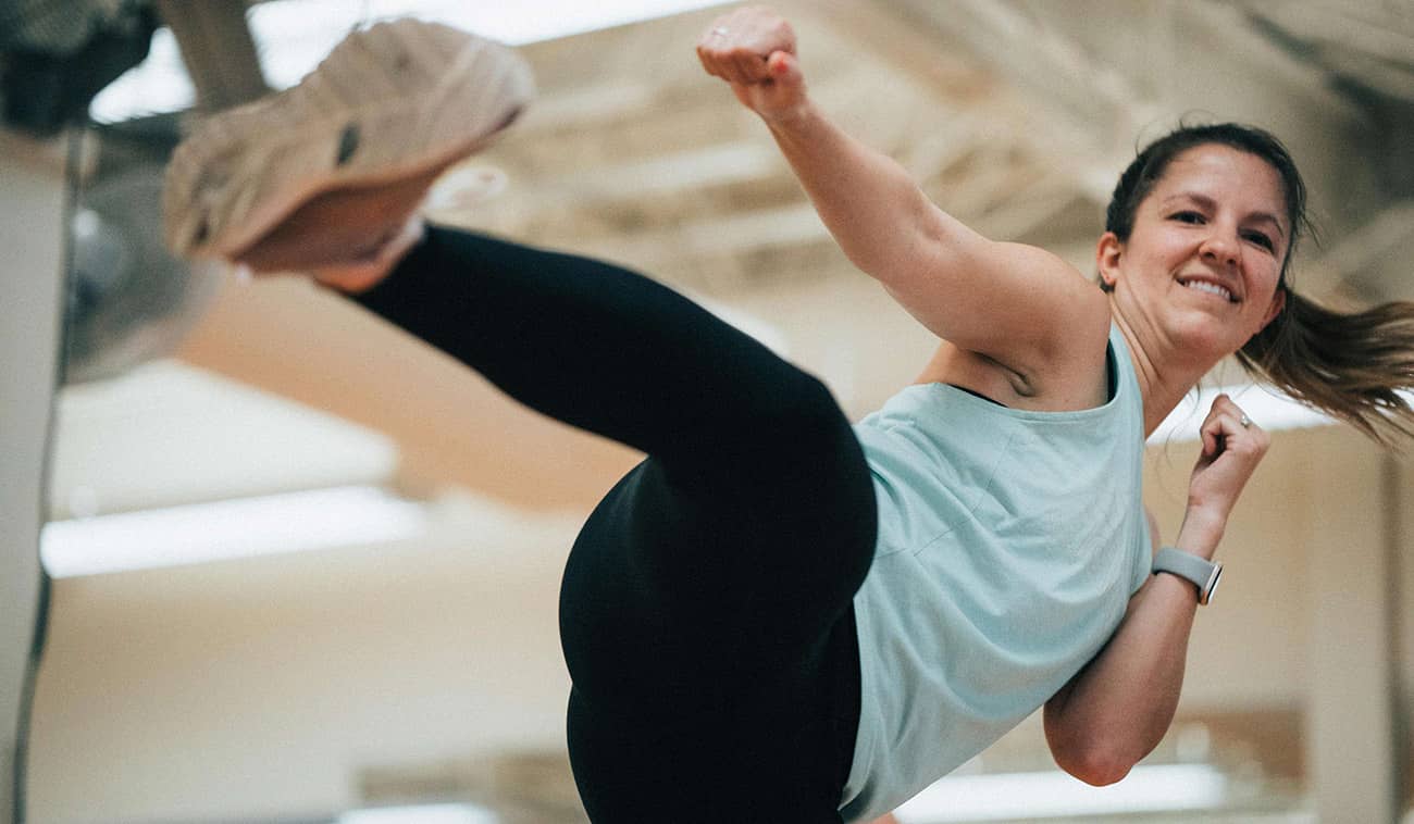 Woman member in blue kicks in the air for her Body Combat class at the TownLake YMCA while smiling for the camera.