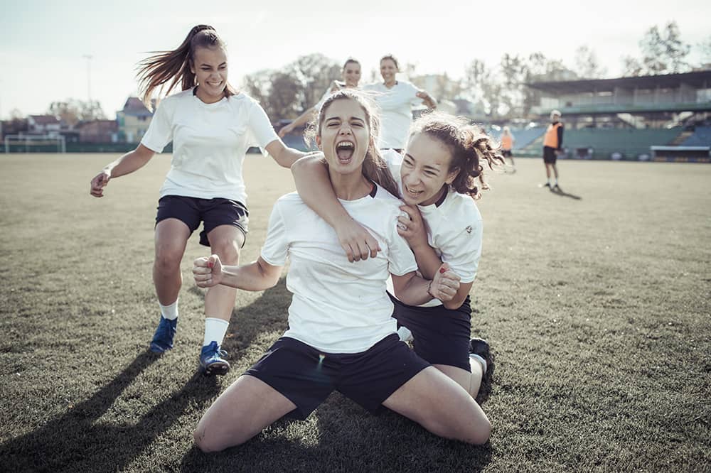 Three teen female soccer players in white jerseys celebrate a scored goal as two other teen female teammates run up behind them to join in the celebration. The teen in focus is looking at the camera, cheering. A grass field is blurred out in the background with the players on the other team, wearing orange soccer jerseys, looking on. 