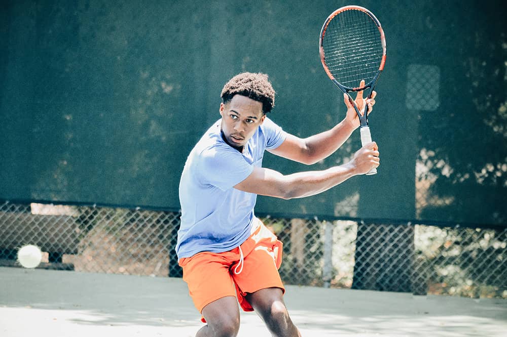 An adult male member in blue and orange holding a tennis racket winds up to hit an incoming tennis ball. A tennis court is blurred out in the background.