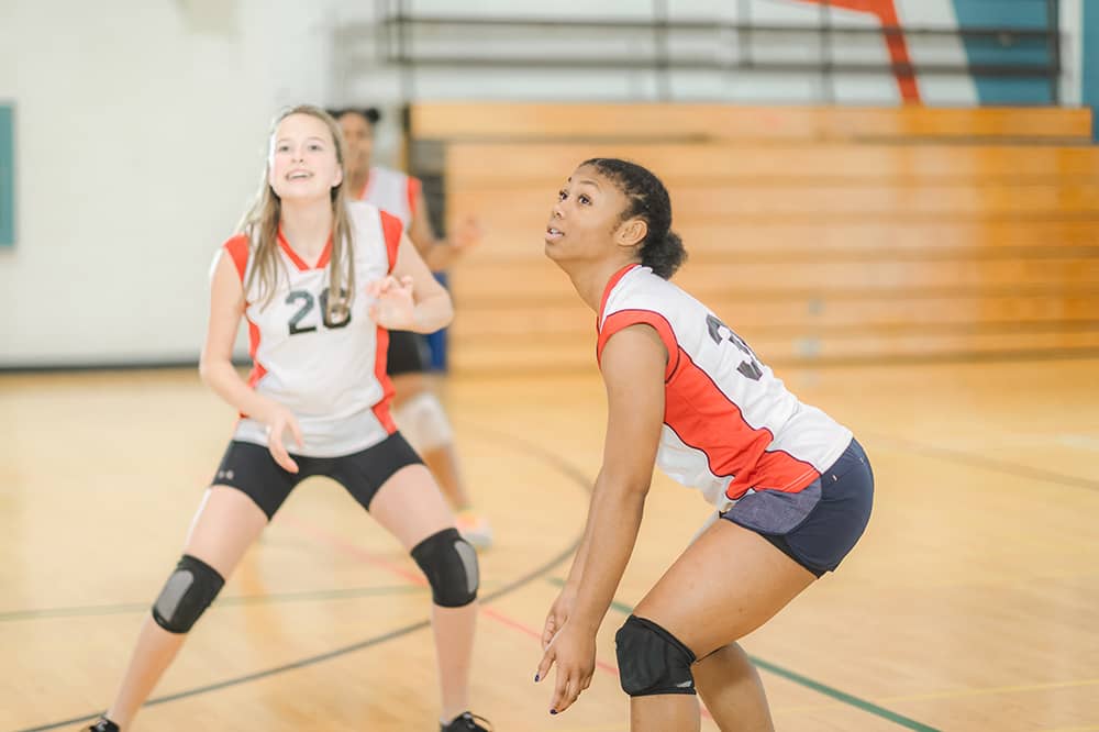 Three teen female members wearing white and red volleyball jerseys smile as they get ready to volley an incoming ball. A basketball gym is blurred out in the background.