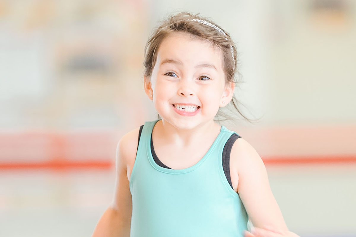 A young girl wearing a blue tank top and black pants runs over a gymnastics mat, smiling. Gymnastics equipment is blurred in the background of the photograph.