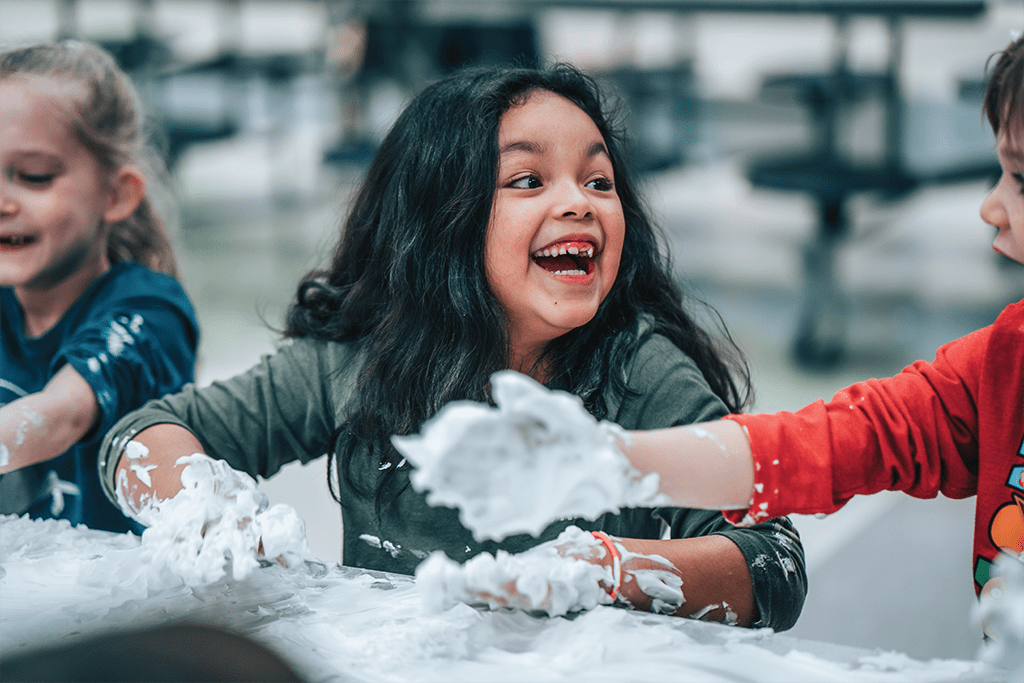 A girl sitting at a table looks to the person on her left, laughing as they play with shaving cream for a fun craft.