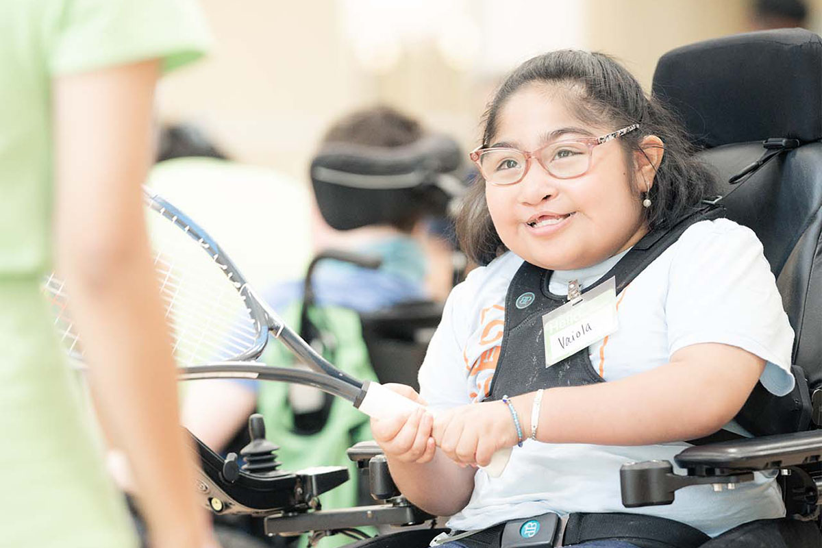 A young camper in a wheelchair wearing a blue shirt and jeans smiles as she holds a tennis racket, looking at a class instructor at Camp In Motion at the YMCA. Other campers in wheelchairs and the walls of the basketball gym are blurred out in the background.