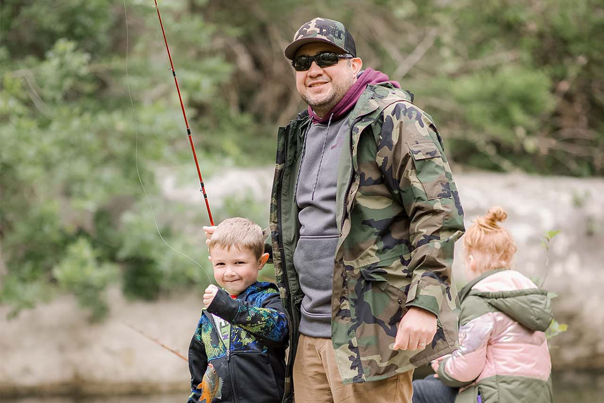 A man in a camo jacket, camo hat, and sunglasses holding a red fishing pole stands next to a boy in a black hoodie holding a fish on the end of the fishing pole’s line. The two are smiling at the camera and standing on a large rock by a creek with other children in jackets fishing behind them. Trees are blurred in the background of the photograph.