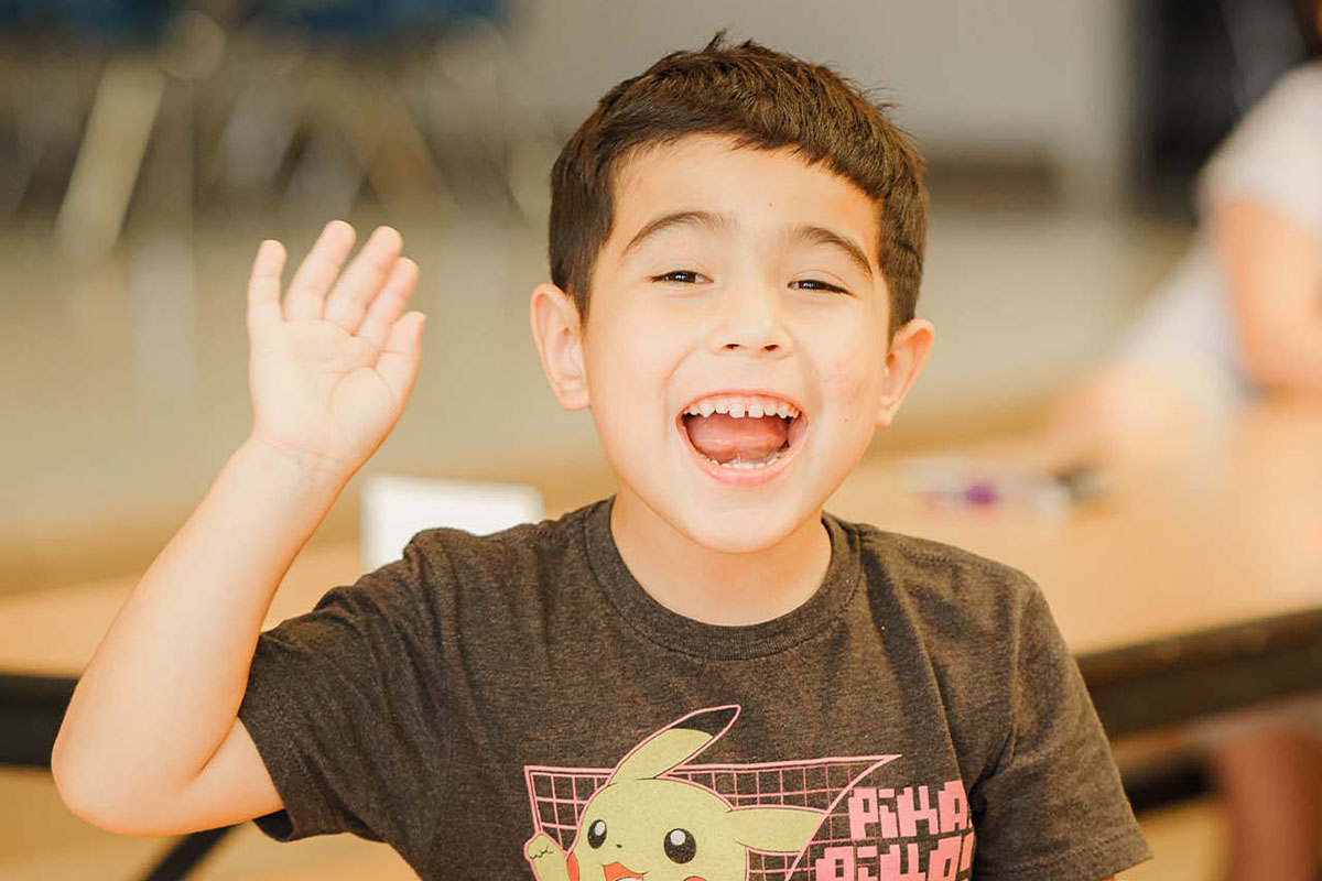 A male camper in a black shirt with building blocks sitting on the cafeteria table in front of him smiles and waves at the camera. Another cafeteria table, chairs, and the walls of a school cafeteria are blurred in the background of the photograph. 