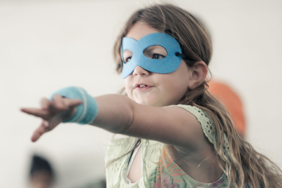 A girl dressed in a green shirt wearing a blue superhero mask and blue cape points and cheers while playing in a branch mini camp activity. Other kids dressed in superhero costumes and the walls of the room they are in are blurred in the background of the photograph.