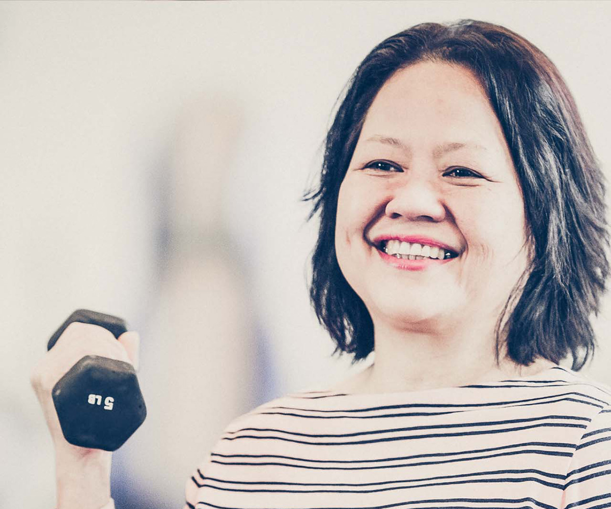 Female adult member in striped shirt sitting in wheelchair lifts a dumbbell and smiles. Weight machines are blurred in the background of the photograph. 