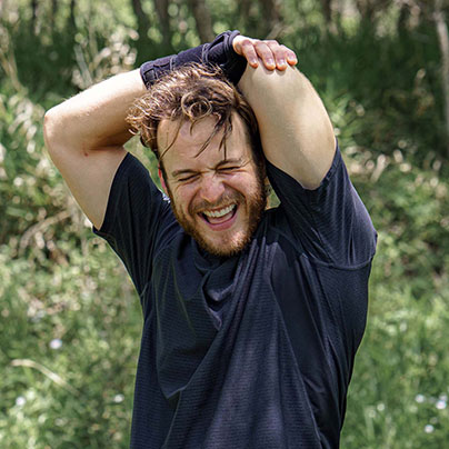 A man stands outdoors and stretches his tricep over his head after exercising. He is smiling and also looks like he has just engaged in strenuous exercise.