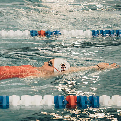 A teen wearing an orange swimsuit,  a white swim cap  and goggles swims a backstroke in a multi-lane pool. Two lanes away, another teen performs a breast stroke.
