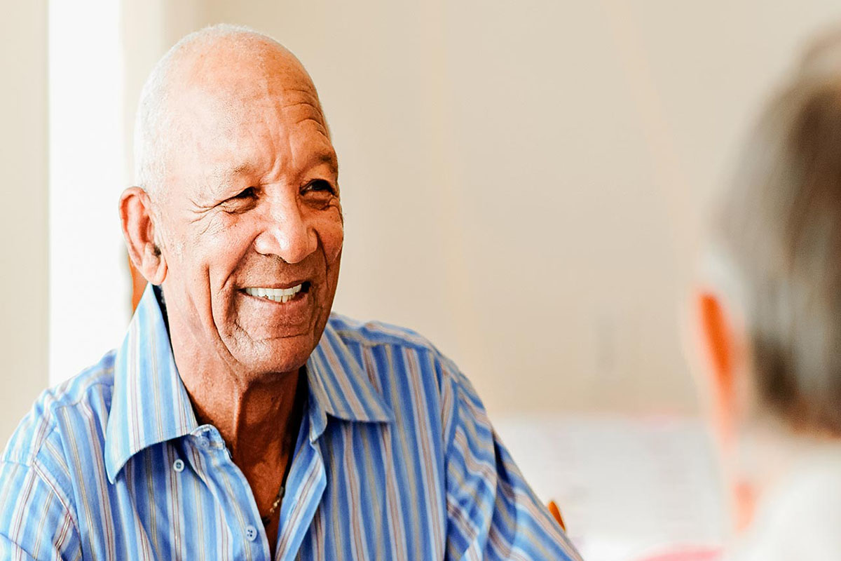 A senior man in a striped blue shirt smiles while seated at a table with other seniors. There is a senior woman to his right in a green shirt and a senior man in front of him in a white shirt. A white wall and window are blurred in the background of the photograph.