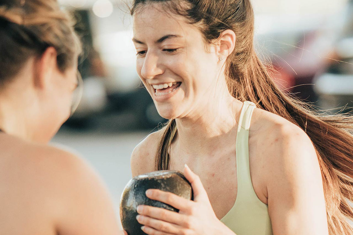 An adult female member in a yellow sports bra laughs as she passes a kettlebell to another adult female member in a black sports bra during a bootcamp class. A parking lot with cars is blurred in the background of the photograph.