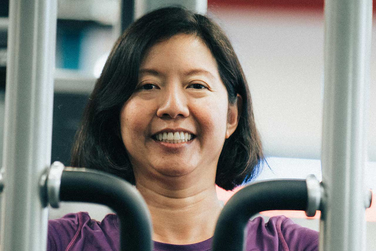 An adult female member in a purple shirt and smiles as she operates a weight machine. Exercise equipment and the wall of the weight room are blurred out in the background of the photograph. 