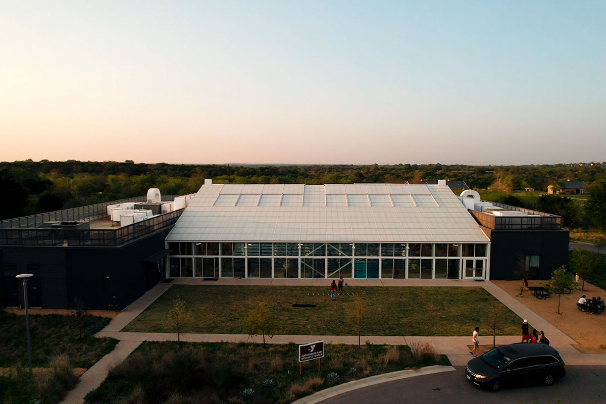 A modern-designed building with a natatorium in the center. Natatorium is white and lined with windows. Buildings on sides are grey and lined with metal. Grass lawn and parking lot sits in front. Behind building is the Camp Moody treeline.