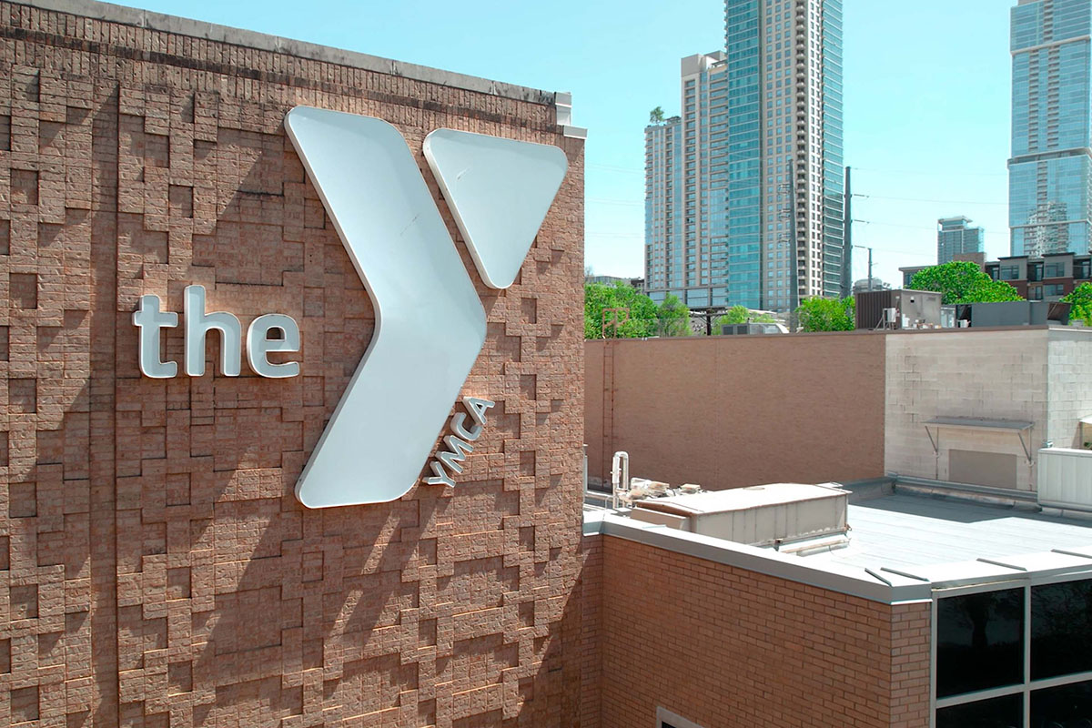 A brick building featuring a prominent white Y logo sits in foreground with part of Austin skyline in background. 