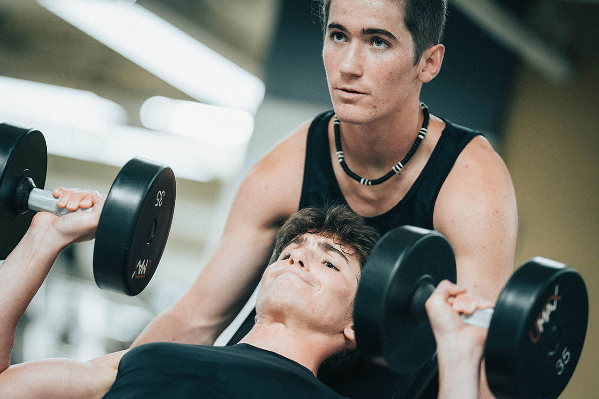 Two young men are lifting weights in partnership. One man is on a weight bench holding heavy dumbbells. His face shows exertion. The other man spots him and looks forward.