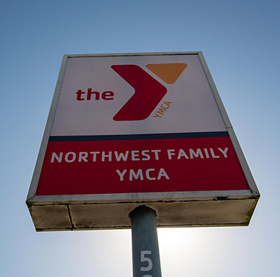 A YMCA location sign is seen from a low angle and features a YMCA logo and reads Northwest Family YMCA. The sky is blue and cloudless behind the sign. 