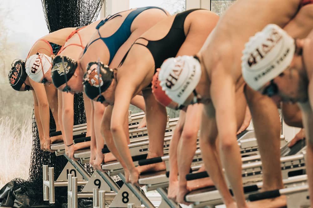 A co-ed line of swimmers stand on diving platforms at the edge of a lap pool. They are bent over and appear ready to dive in for a race.