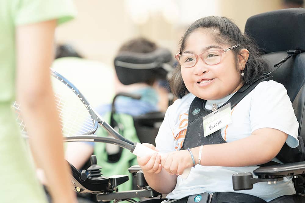 A young girl in a wheelchair wearing a blue shirt and jeans smiles as she holds a tennis racket, looking at a class instructor at Camp In Motion at the Southwest Family YMCA. Other children in wheelchairs and the walls of the basketball gym are blurred out in the background.