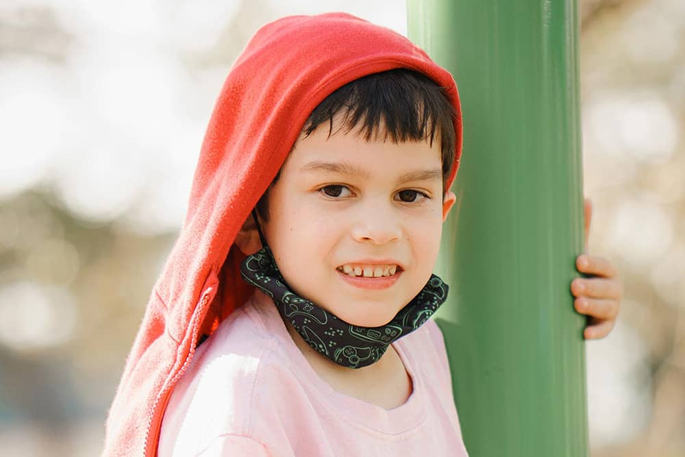 A young boy in a pink shirt with a red hoodie draped over his head smiles at the camera while seated on a green bar on a playground. Trees are blurred in the background of the photograph. 