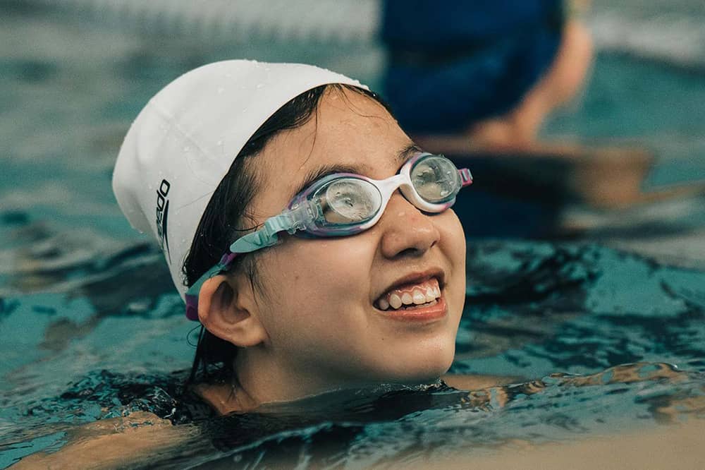 A girl in a white swim cap and goggles looks up at her swim instructor off camera as she treads water in a lap lane. Another child in a blue swim cap and lap lanes are blurred in the background of the photograph.