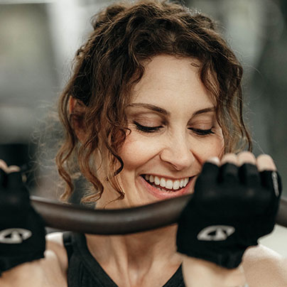 A woman wearing black weightlifting gloves holds a curved barbell. She smiles as she flexes the barbell.
