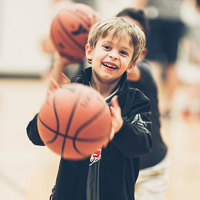 A smiling boy wearing a black jacket and red shorts is shooting a basketball. The ball is blurry, showing motion.