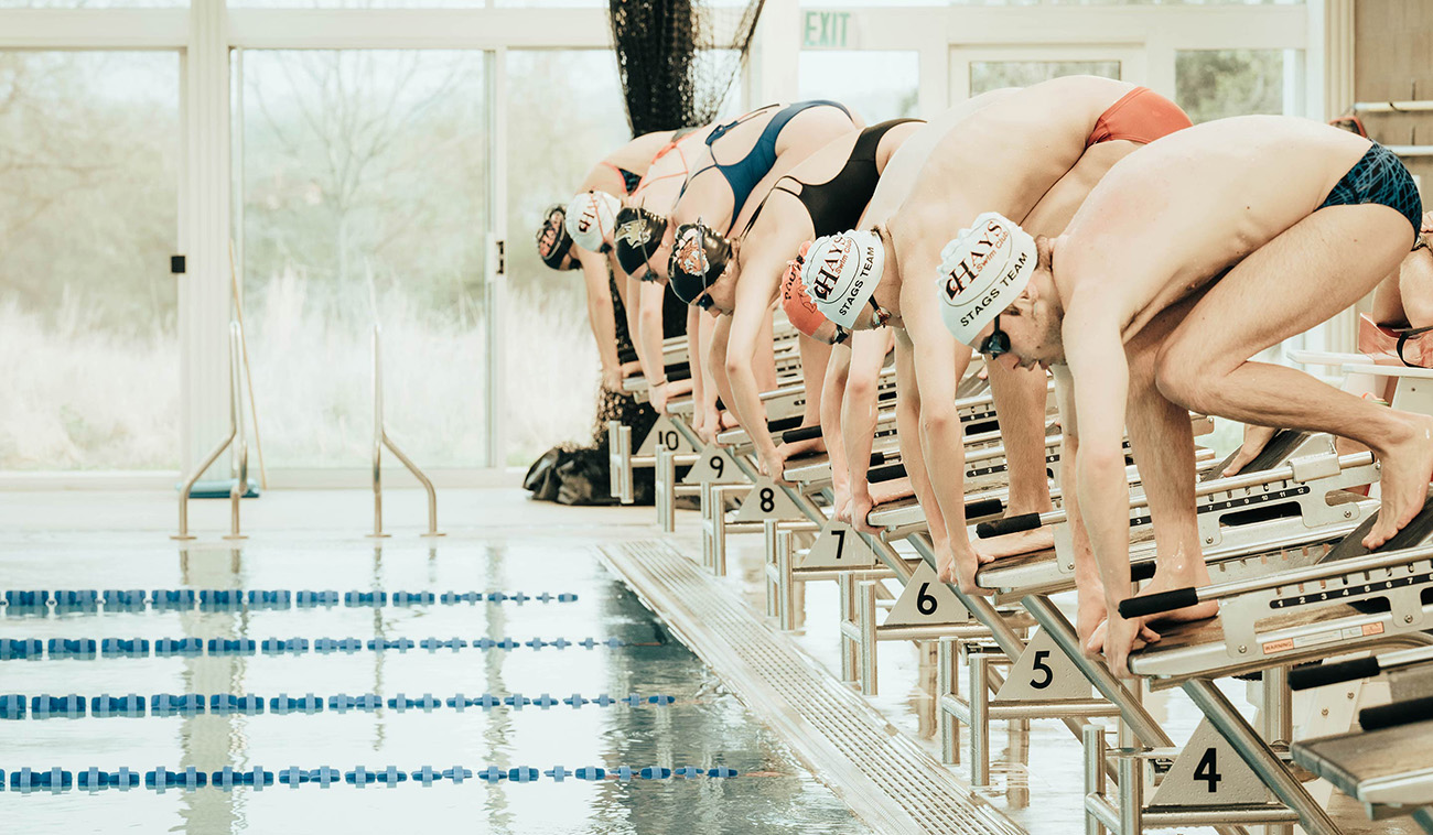 Seven young adult swimmers in goggles and swim caps get ready to dive off of their swimming blocks at YMCA Camp Moody. Pool lanes and the natatorium’s windows are blurred in the background of the photograph. 