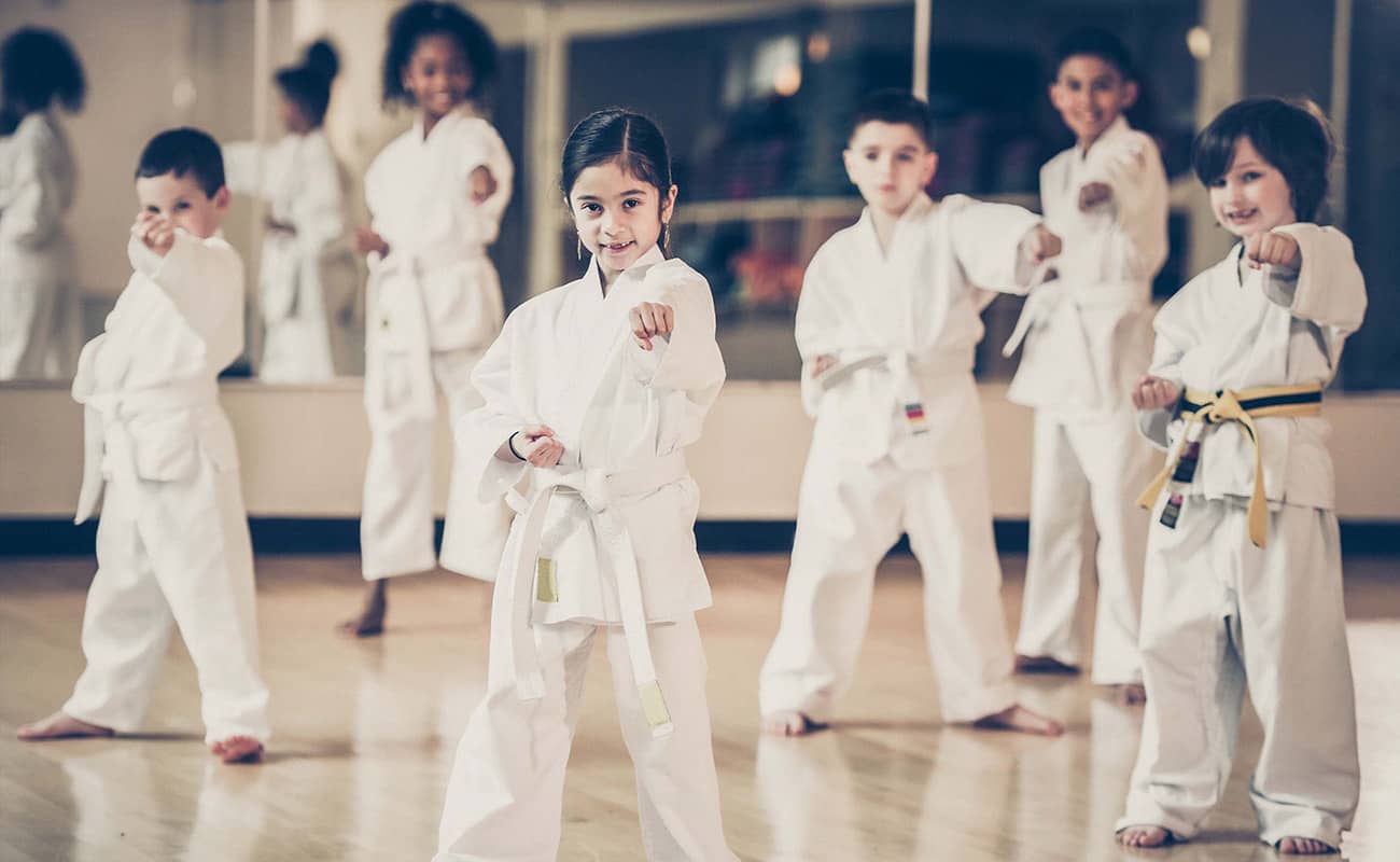 Six children members, two girls and three boys, in Taekwondo uniforms throw a punch and smile for the camera. An empty workout room and wall mirror are blurred in the background of the photograph.