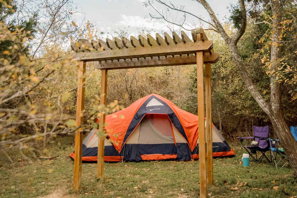 An orange, blue, and tan colored tent sits in a campsite with purple and blue lawn chairs to the right of the tent and a wood arbor in front of it. Trees with autumn-colored leaves are blurred in the background of the photograph. 