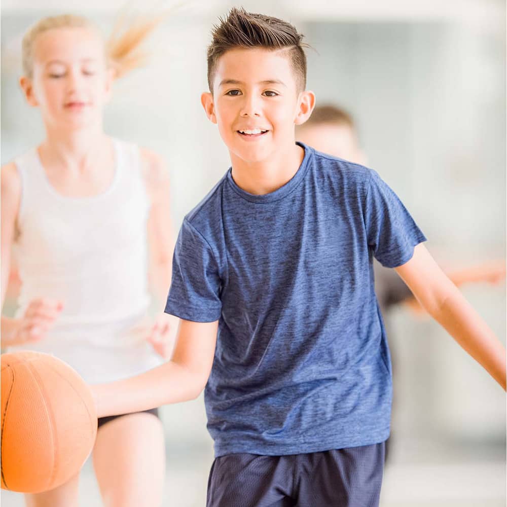 A boy in a blue shirt smiles as he dribbles a basketball in a basketball gym. A girl in a white sleeveless shirt and the walls of the basketball gym are blurred in the background of the photograph.