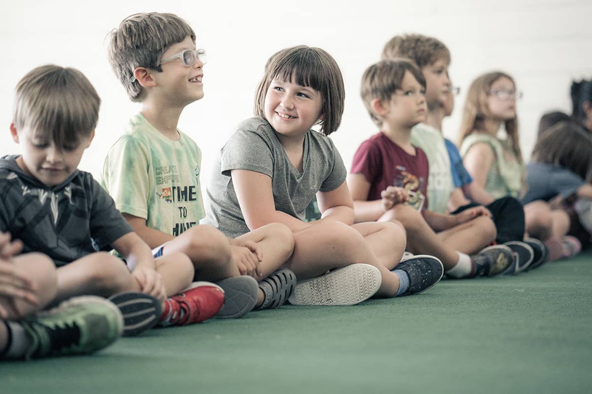 A girl in a gray shirt and a boy in a green shirt and glasses share a smile together while seated on the floor in a line with a group of other kids. The green floor and white walls of the room they are in are blurred in the background of the photograph. 