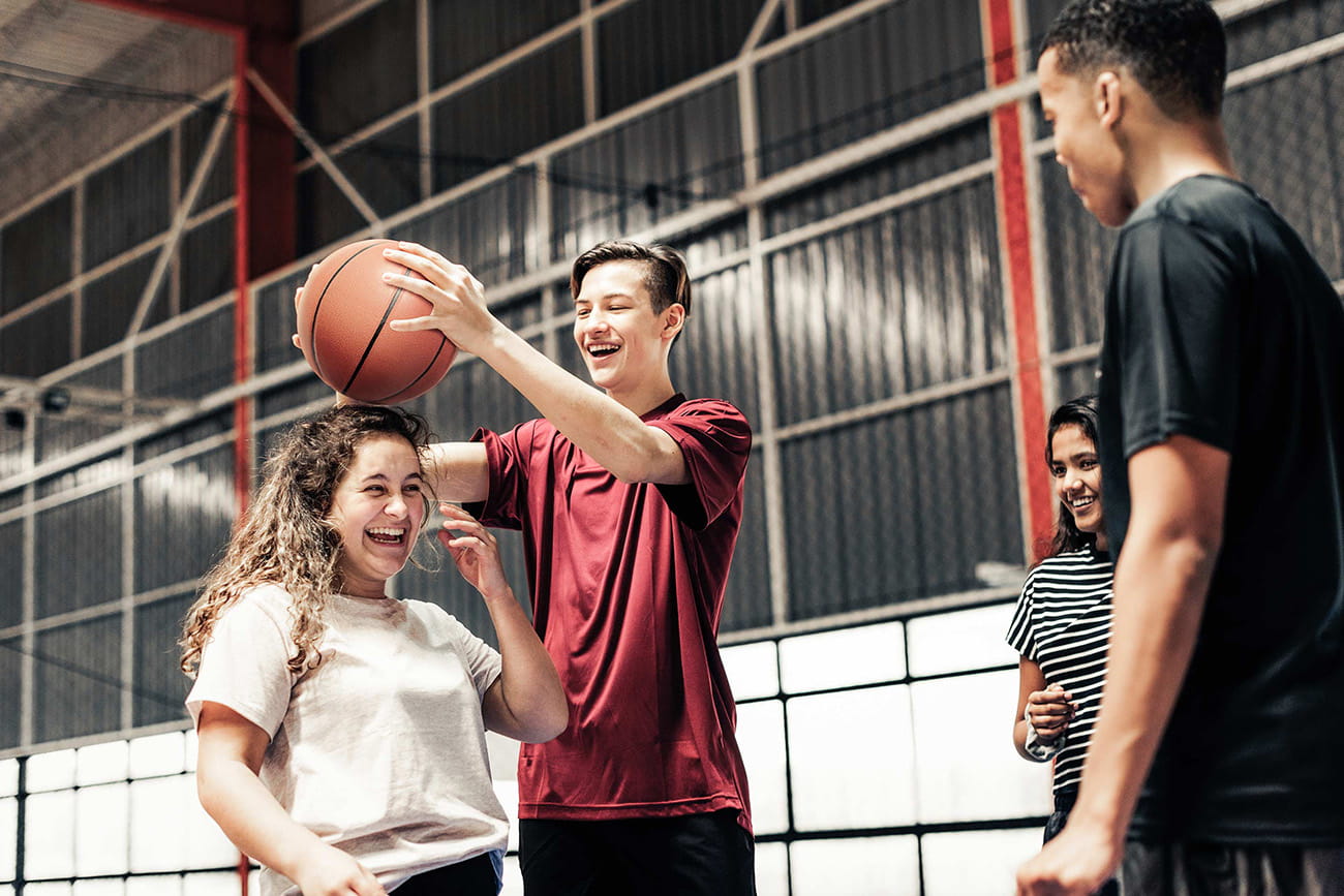 A male teen in a red shirt playfully sets a basketball on top of a teen female’s head as they laugh together. A male teen in a black shirt and a female teen in a striped shirt smile and watch on. A basketball gym is blurred in the background of the photograph. 
