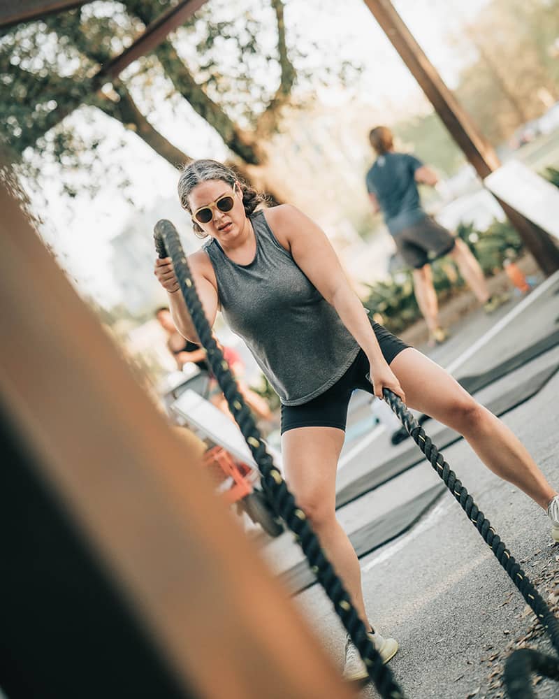 An adult female member in a gray sleeveless shirt pulls on black battle ropes during an outdoor bootcamp class. Steel beams, yoga mats, and a classmate are blurred in the background of the photograph. 