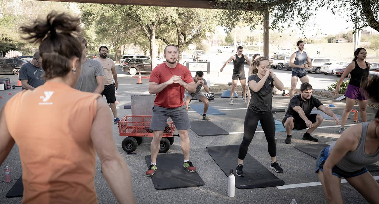 A large group of adult members in workout clothes do squats in a parking lot during a bootcamp class. Yoga mats and water bottles lay on the ground in front of them. A parking lot filled with cars is in the background of the photograph.