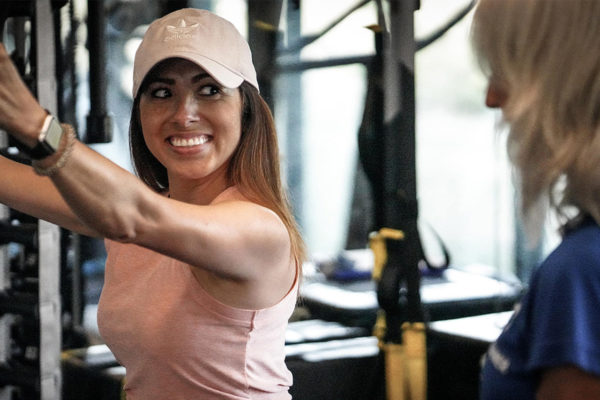 An adult female member in a pink sleeveless shirt and pink hat smiles as she listens to her female adult trainer in a blue shirt give directions on how to correctly operate a weight machine. Weight room equipment is blurred out in the background of the photograph. 