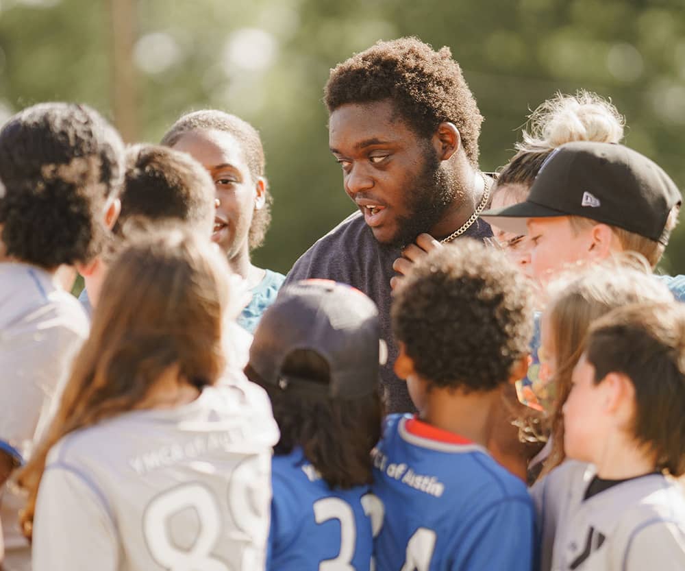 A volunteer coach in a gray shirt speaks in a huddle of kids wearing gray and blue soccer jerseys. A soccer field and trees are blurred in the background of the photograph. 