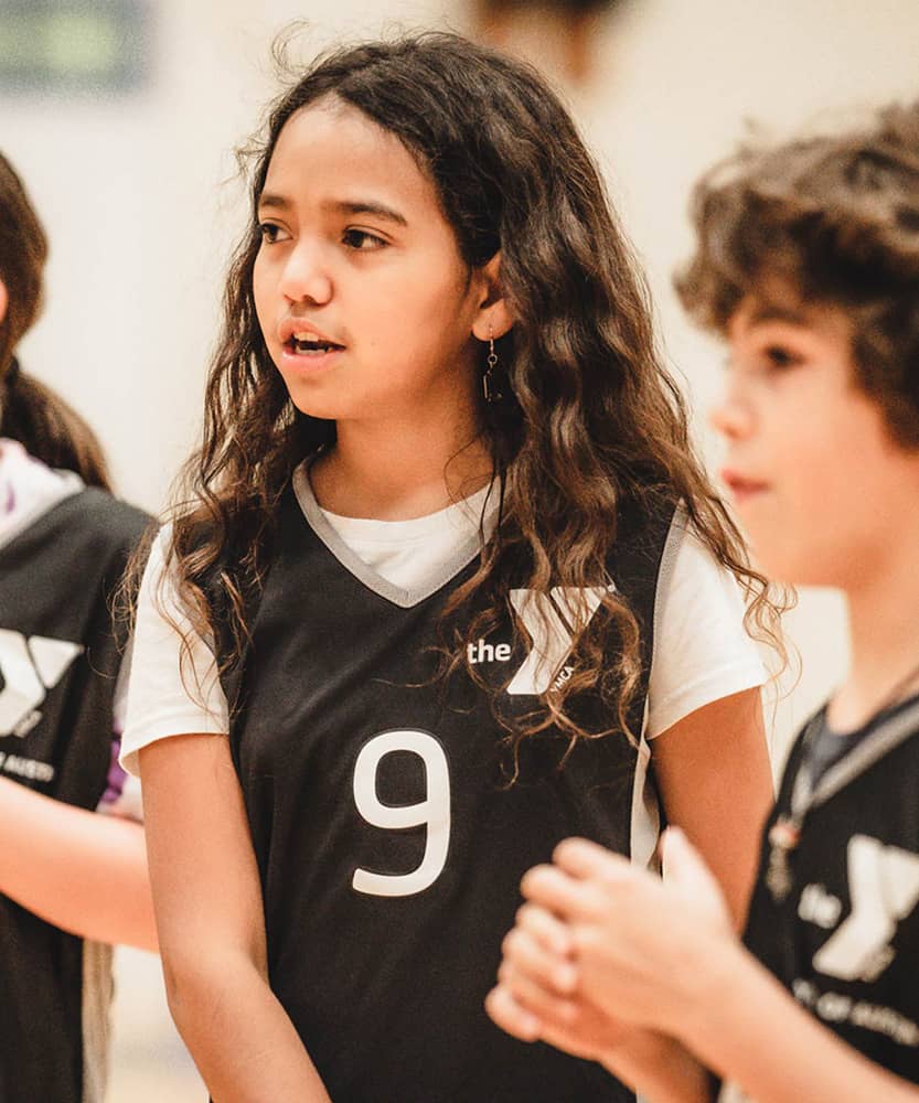 A group of two girls and one boy wearing black basketball jerseys chat between drills of a basketball sports clinic. A basketball gym is blurred in the background of the photograph. 