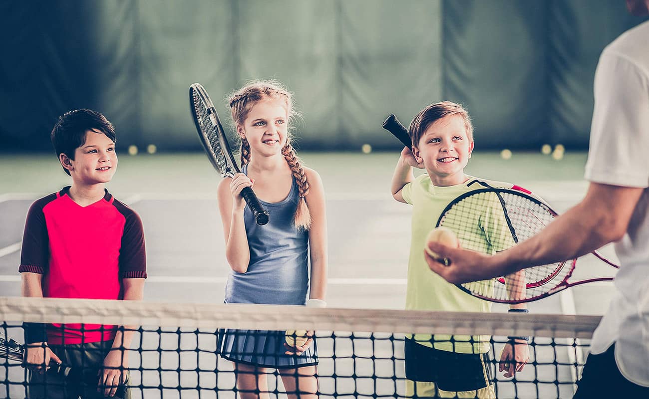 Two boys and one girl, dressed in red, blue, and green shirts, holding tennis rackets, listen to their male instructor in white teach them tennis skills. The instructor is standing on one side of the net and the kids are standing on the other, facing the camera. A tennis court and tennis balls are blurred in the background of the photograph.