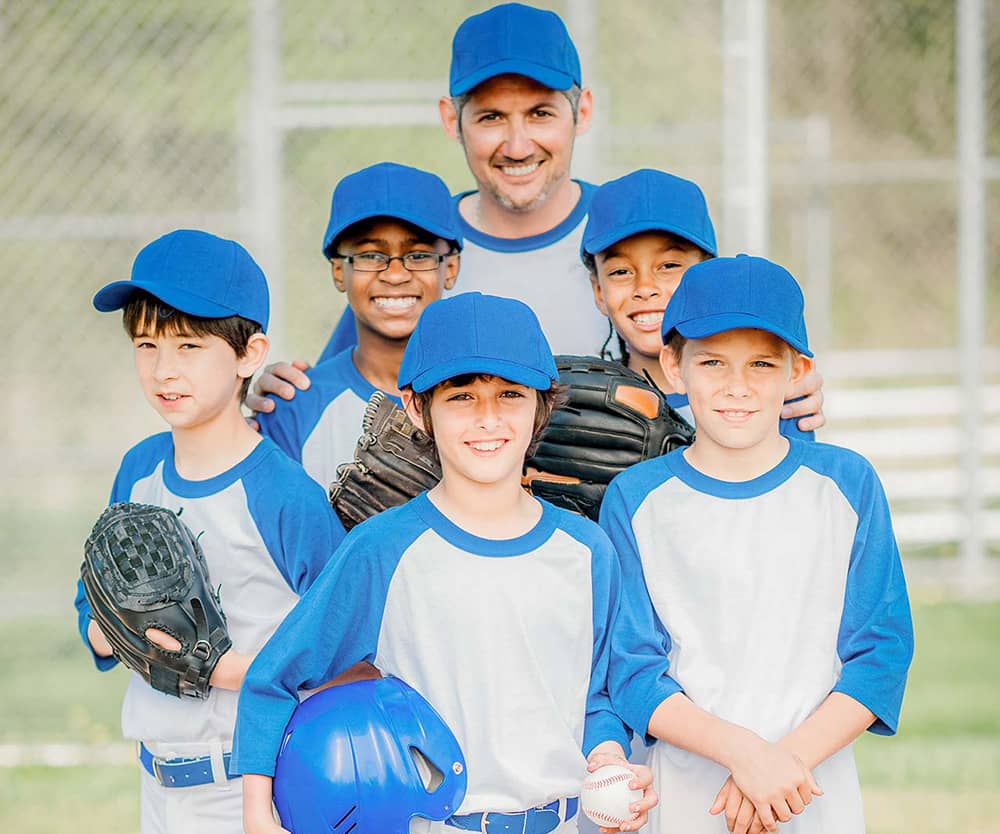 Five boys and a male volunteer coach dressed in blue and white baseball uniforms stand together on home plate and smile for the camera. A dugout fence is blurred in the background of the photograph. 