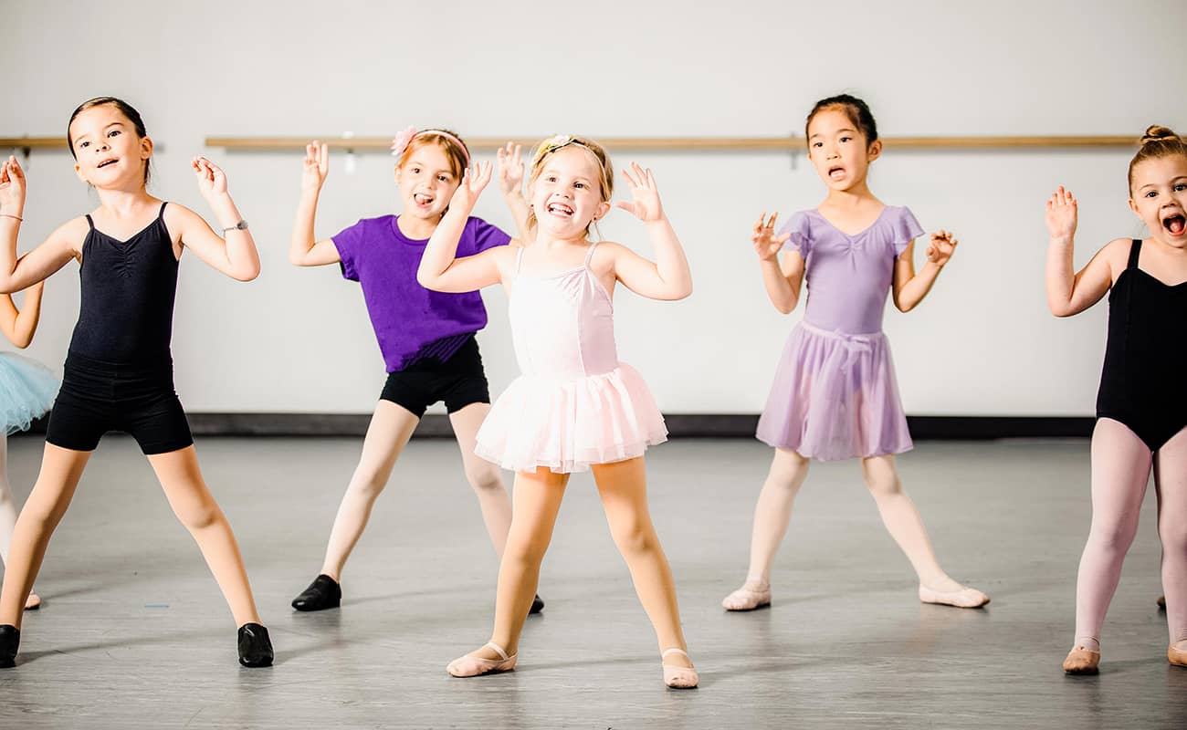 Six girls wearing different colored leotards and tutus look up to their dance instructor and while doing the chicken dance. A dance studio is blurred in the background of the photograph. 