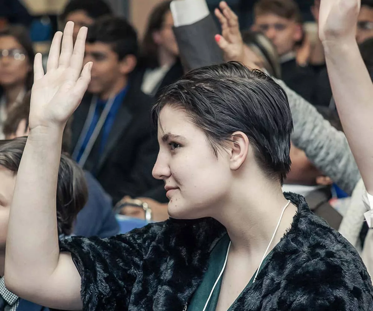 A female YG teen dressed in business professional attire raises her hand while seated during a District Conference ceremony in a high school auditorium. Seated teens and the walls of the auditorium are blurred in the background.