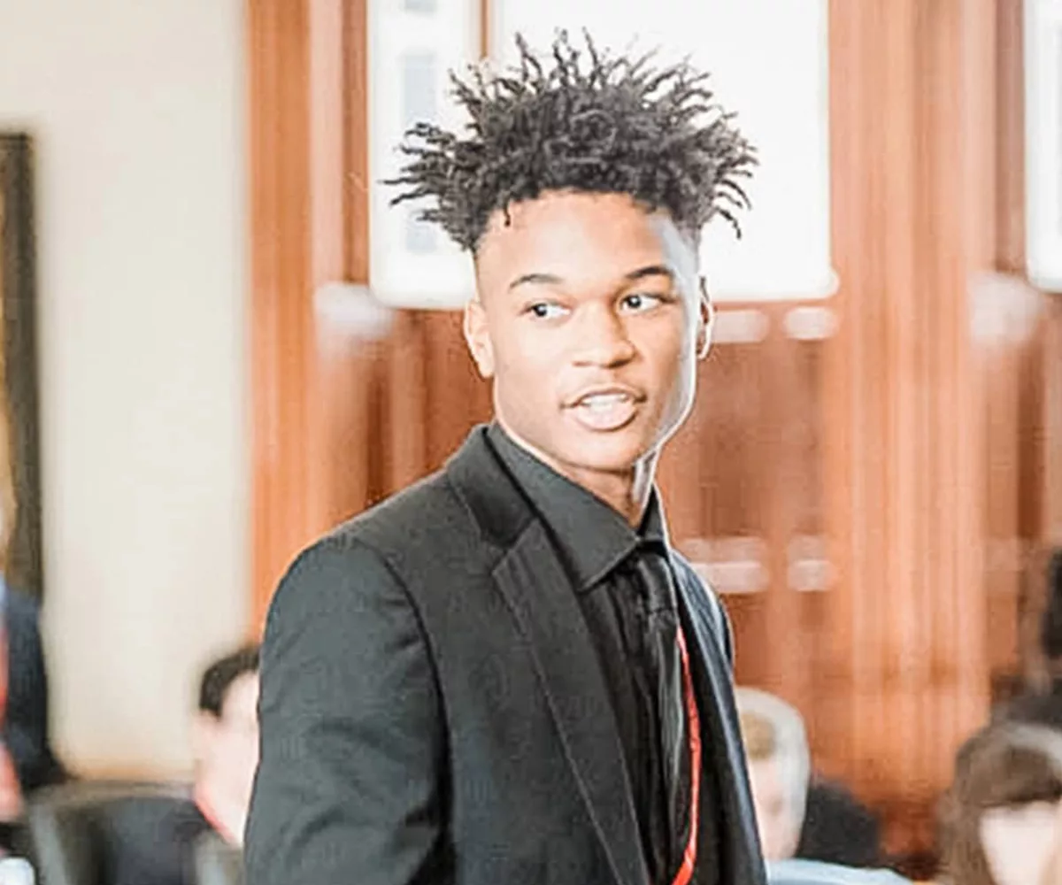 A male YG teen dressed in business professional attire stands and speaks to his peers in the Texas Capitol Senate Chamber. Other teens dressed in business professional attire seated at desks and the walls of the Texas Capitol Senate Chamber are blurred in the background of the photograph.