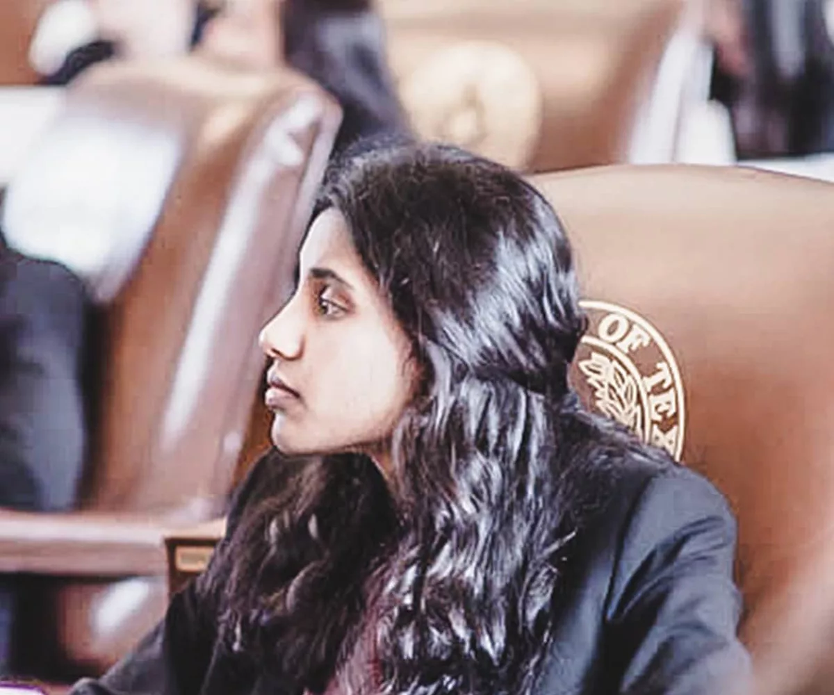 female YG teen dressed in business professional attire listens to her colleagues while sitting at a desk in the Texas Capitol Senate Chamber. Other teens dressed in business professional attire seated at desks are blurred in the background of the photograph.