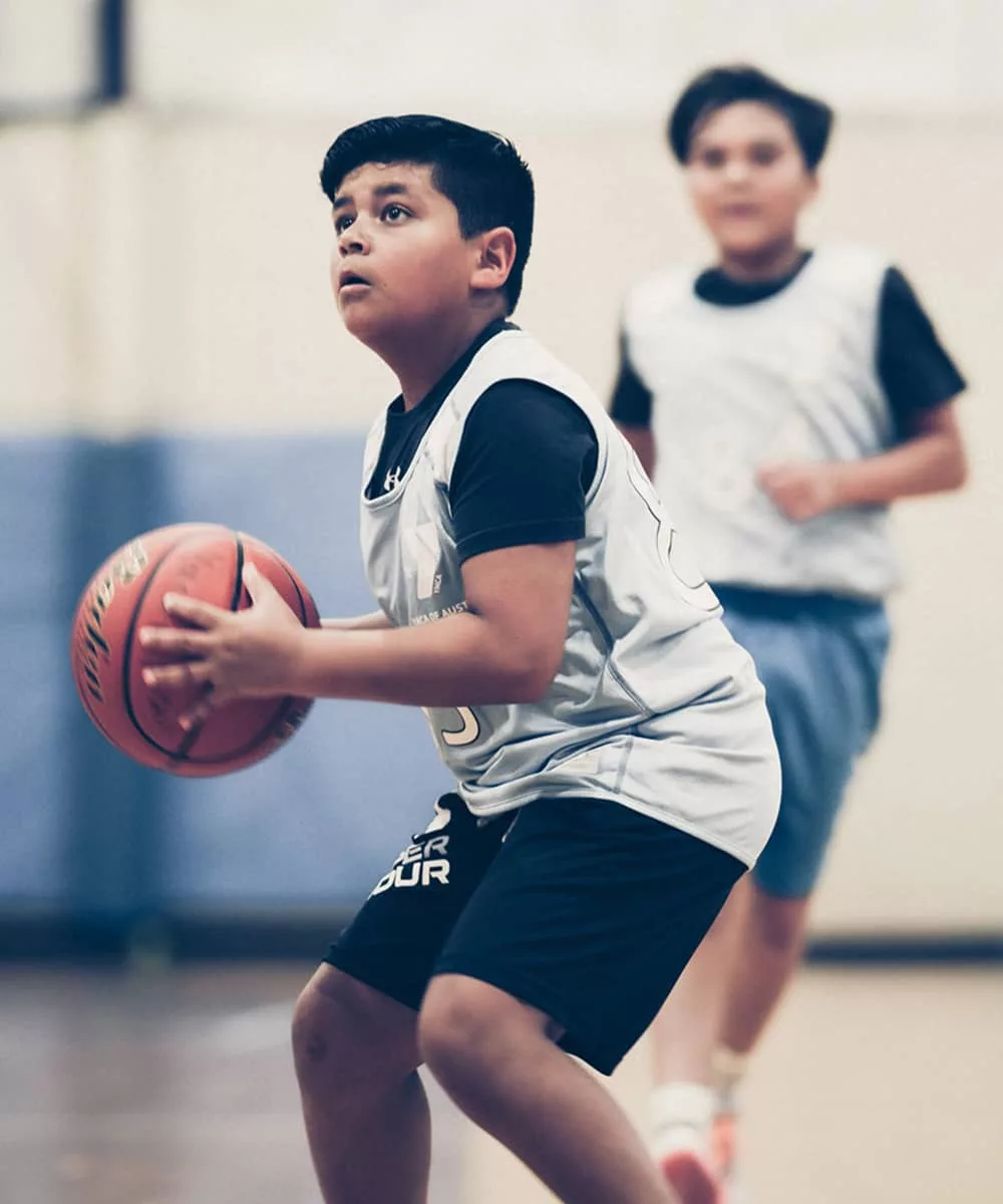 A boy in a blue basketball uniform prepares to shoot a jump shot during a basketball game as a teammate in a blue basketball uniform behind him watches on. A basketball court is blurred in the background of the photograph.