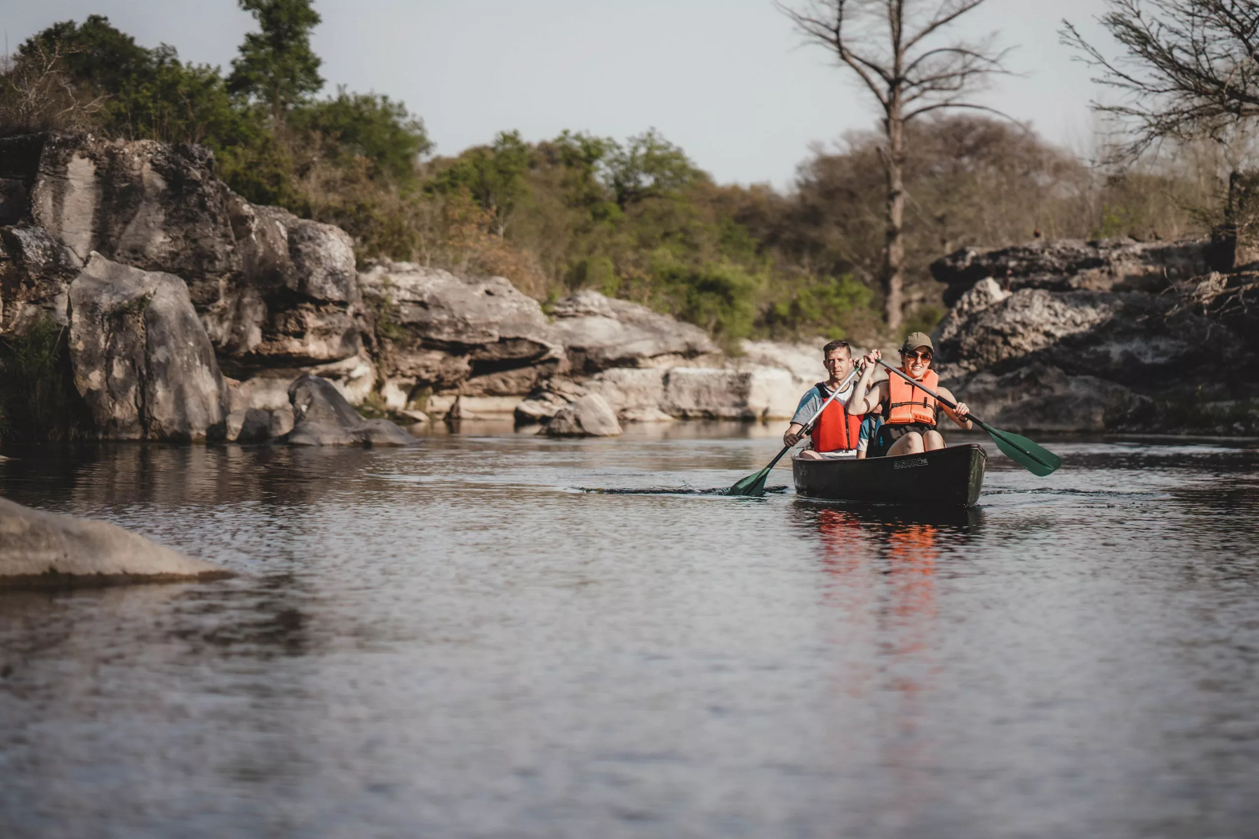 Two people are in a canoe with their oars in the water. They are wearing orange lifejackets. A rocky outcropping is behind them.