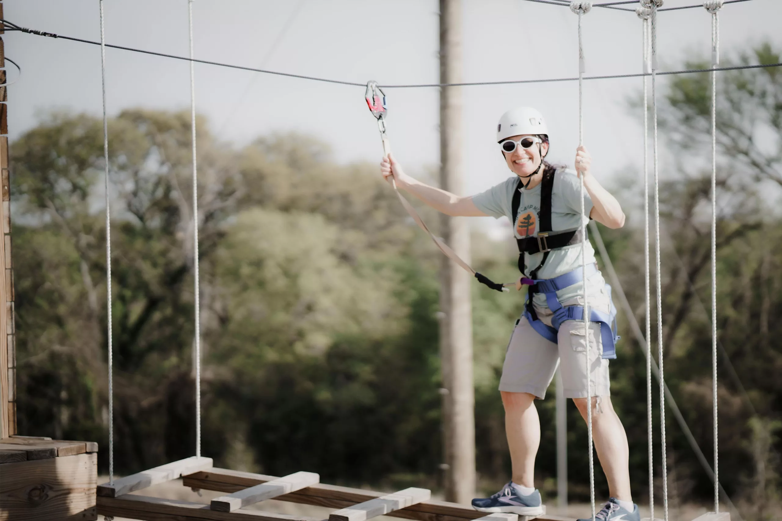 A woman wearing a safety helmet and sunglasses smiles as she navigates a high ropes course.