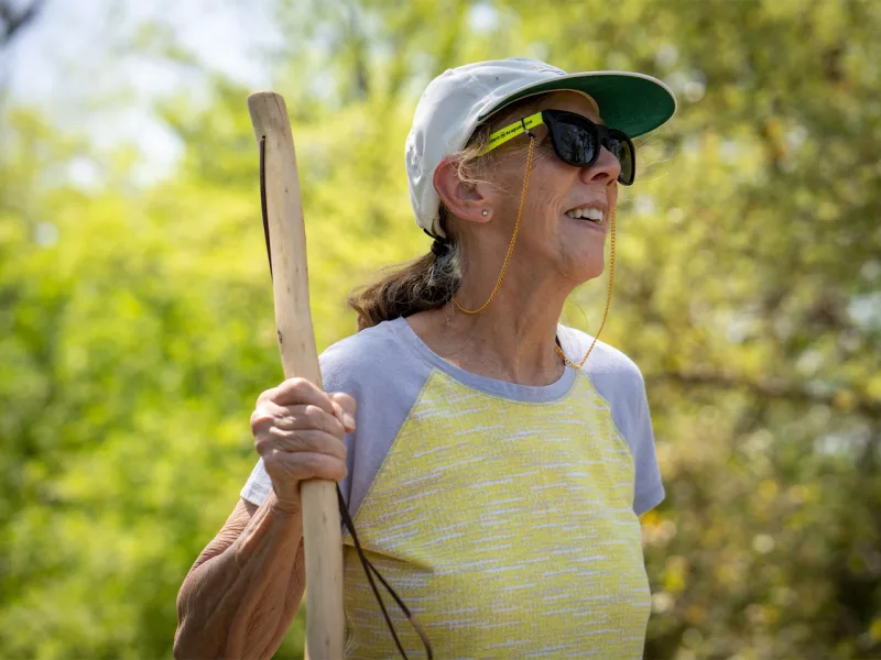 A woman wearing sun shades and a hat walks in nature using a walking stick