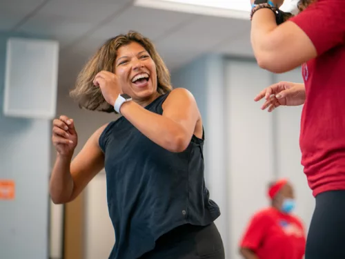 A woman smiles while enthusiastically dancing and looks to another dancer in a group fitness dance class.