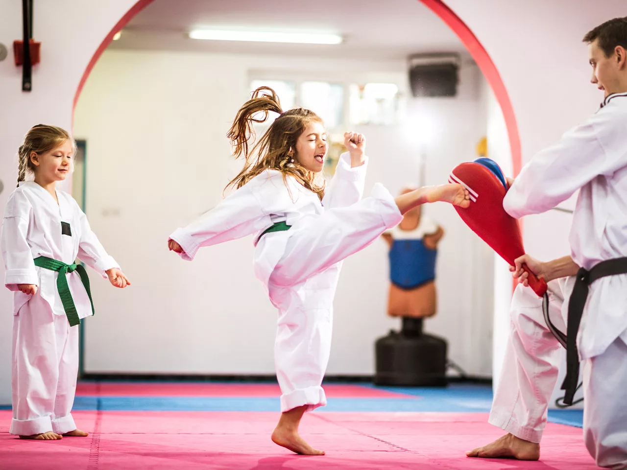 A child wearing a martial arts uniform performs a kick. Her instructor is holding a red foam board.