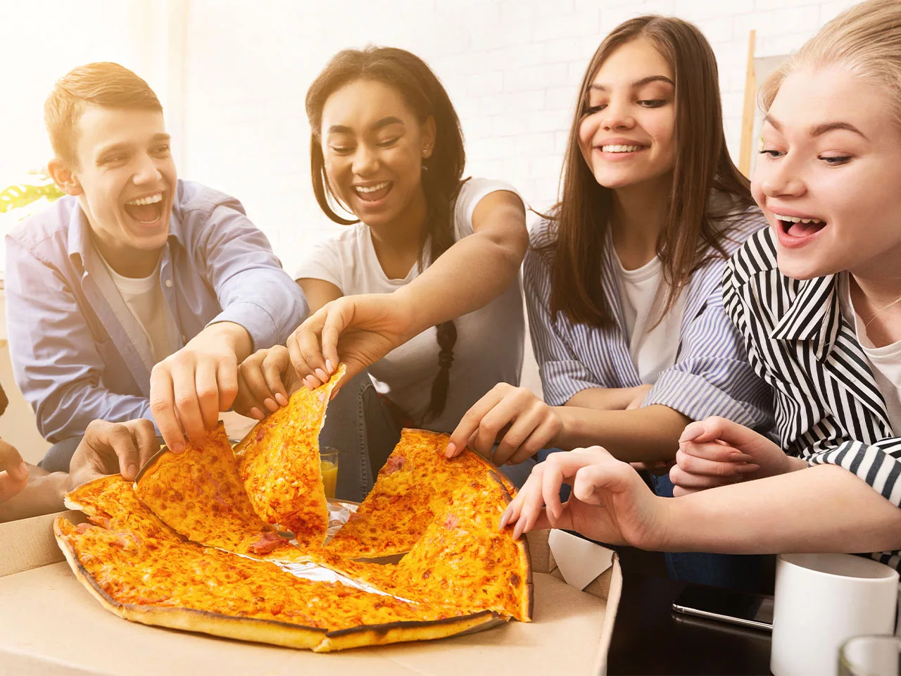 Teens sit around a large cheese pizza, each smiling and taking a slice.